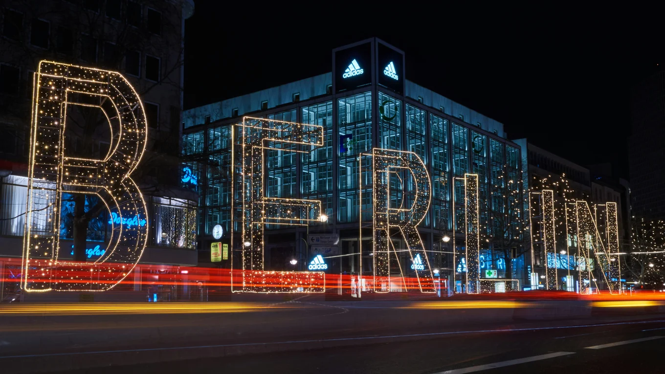 A dark city street at night with a big square building lit up in the background. Following the street from left to right the name 'Berlin' is written in large, illuminated letters.