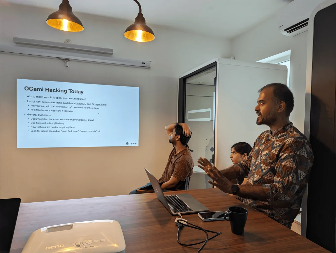 KC is sitting on a bar stool with his laptop open in front of him on the right hand side of the image. He is speaking and making a gesture with his hands, looking to his right towards his presentation slide. The slide is at the centre of the image, and it says OCaml Hacking Today with some smaller bullet points beneath the heading. There are two other people in the image also looking at the presentation slide, both sitting on chairs on the right hand side behind KC.