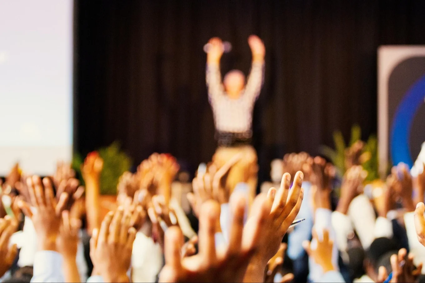 Blurry speaker, audience with hands raised, indoors.