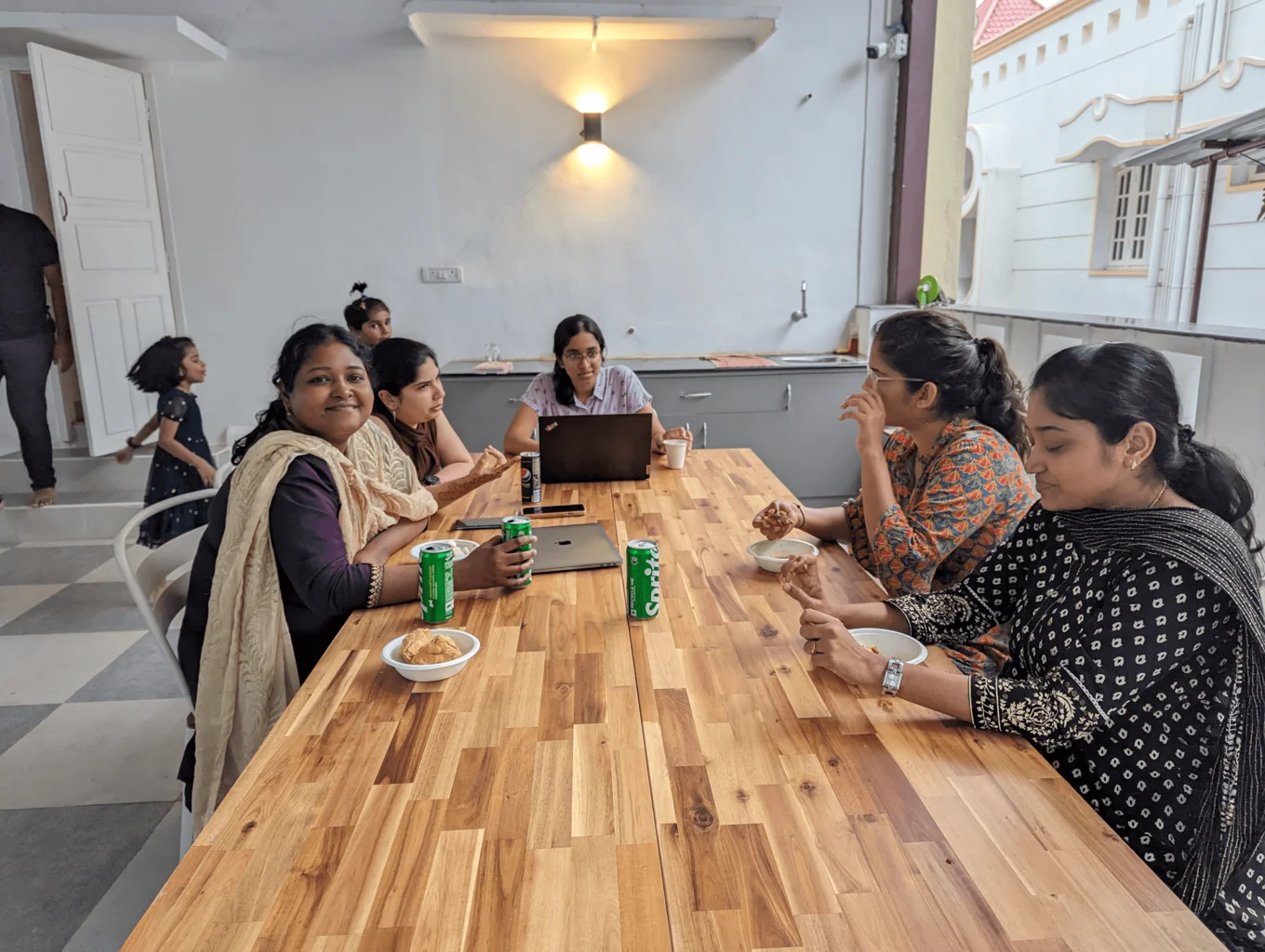 Five women are seated around a dining table on the terrace having food. The angle is from head of the table looking at the other end. One woman is looking into the camera and smiling. In the background are two young children playing. The wall behind the table is white, and to the right is a banister behind which is the white wall of another house.