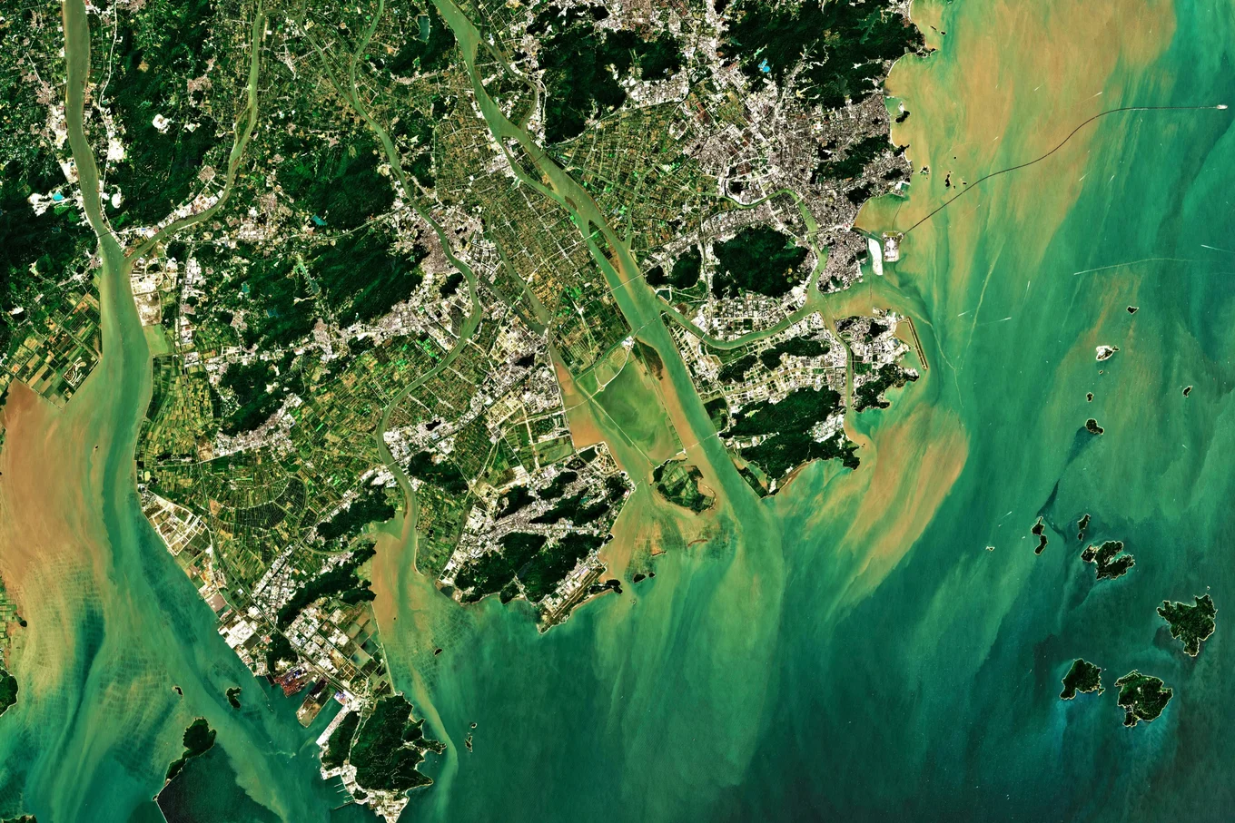 A satellite view of a river delta where green farmland and a dense city meet turquoise and sediment-brown coastal water.