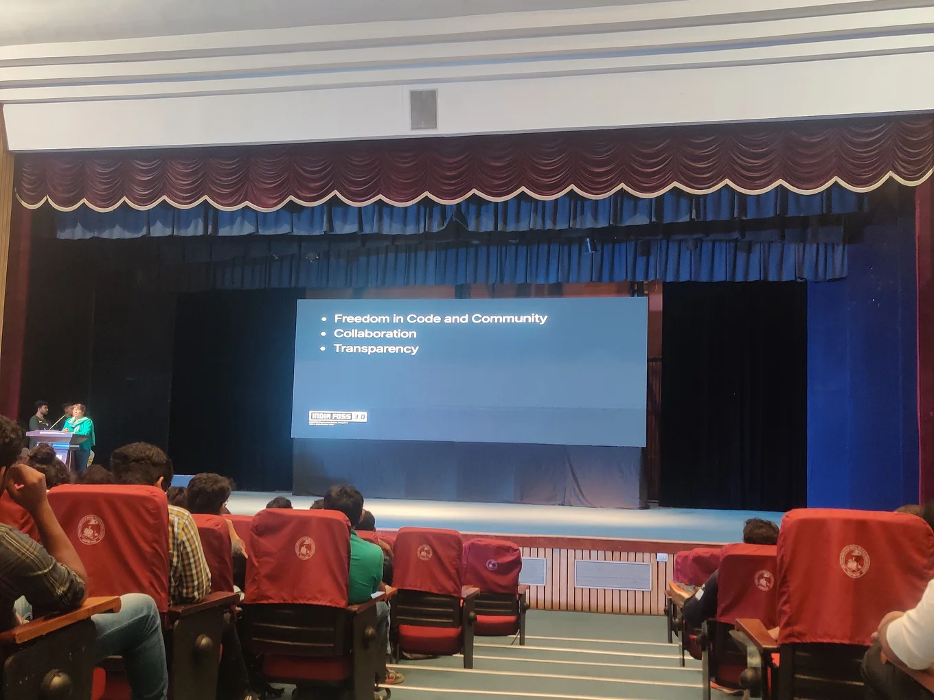 An auditorium with a stage and red and blue curtains surrounding it. A presentation slide is projected onto the centre-back wall of the stage. It has three bullet points which read: freedom in code and community, collaboration, transparency. There are people in red chairs facing the stage and paying attention to the talk.