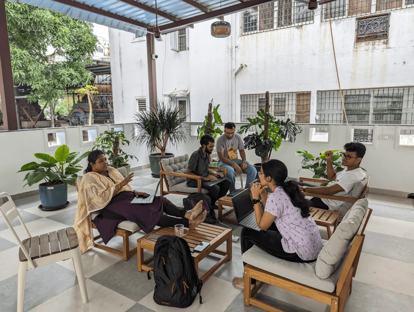 Five people are sitting outside on a large covered roof terrace. They are gathered around two coffee tables with their laptops. Three of them are having a discussion, and the two others are looking at a laptop and focusing on that. There are several potted plants along the railing of the terrace, and we can see the wall of a white house in the background as well as a tree.