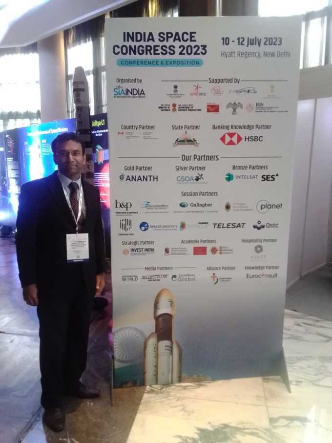 Shakthi is standing next to a vertical sign from the India Space Congress 2023 conferece. The sign features the logos of partners and sponsors.