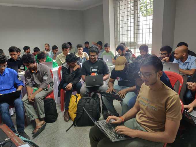 A group of people gathered in a room. They're sitting on chairs in a half-circle formation facing the camera. Most are intently looking at either their own or their neighbour's laptop.