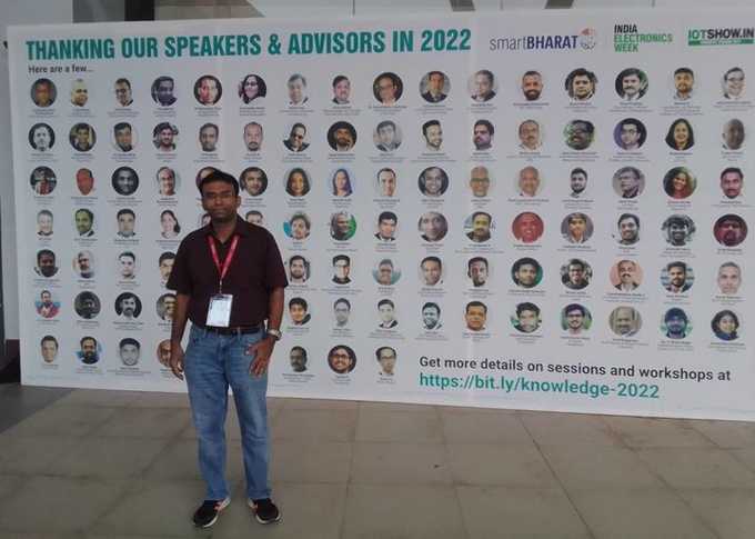 Shakthi is standing in front of a sign which says "thank you to our speakers & advisors in 2022", the sign has headshots of different speakers at the conference.