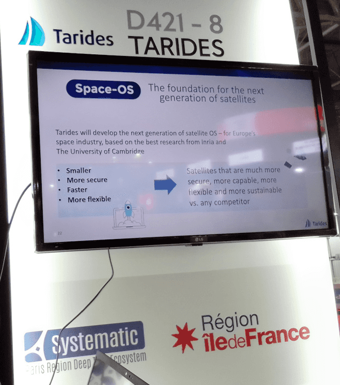 A monitor showing a slide which says SpaceOS - the foundation for the next generation of satellites. Tarides will develop the next generation of satellite OS – for Europe's space industry, based on the best research from Inria and The University of Cambridge. Bullet points: smaller, more secure, faster, more flexible. Arrow pointing to: Satellites are much more secure, more capable, more flexible and more sustainable vs. any competitor. The monitor itself is on a wall with the logos of Tarides, Systematic, and Région Île de France